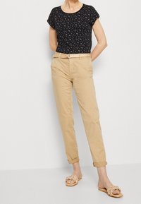 Svart kortärmad topp med blommönster, beige upprullade chinos, flätad beige bälte och ljusbruna slip-on sandaler med texturerade remmar.