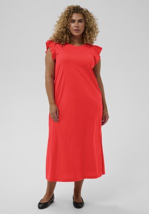 Göndör hajú nő piros ujjatlan midi ruhában, fodros vállrésszel és fekete lapos cipőben, tiszta háttér előtt állva.