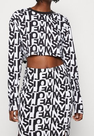 Person iført sort og hvid crop top og nederdel med gentaget "TOMMY JEANS" tekstmønster og lange ærmer.