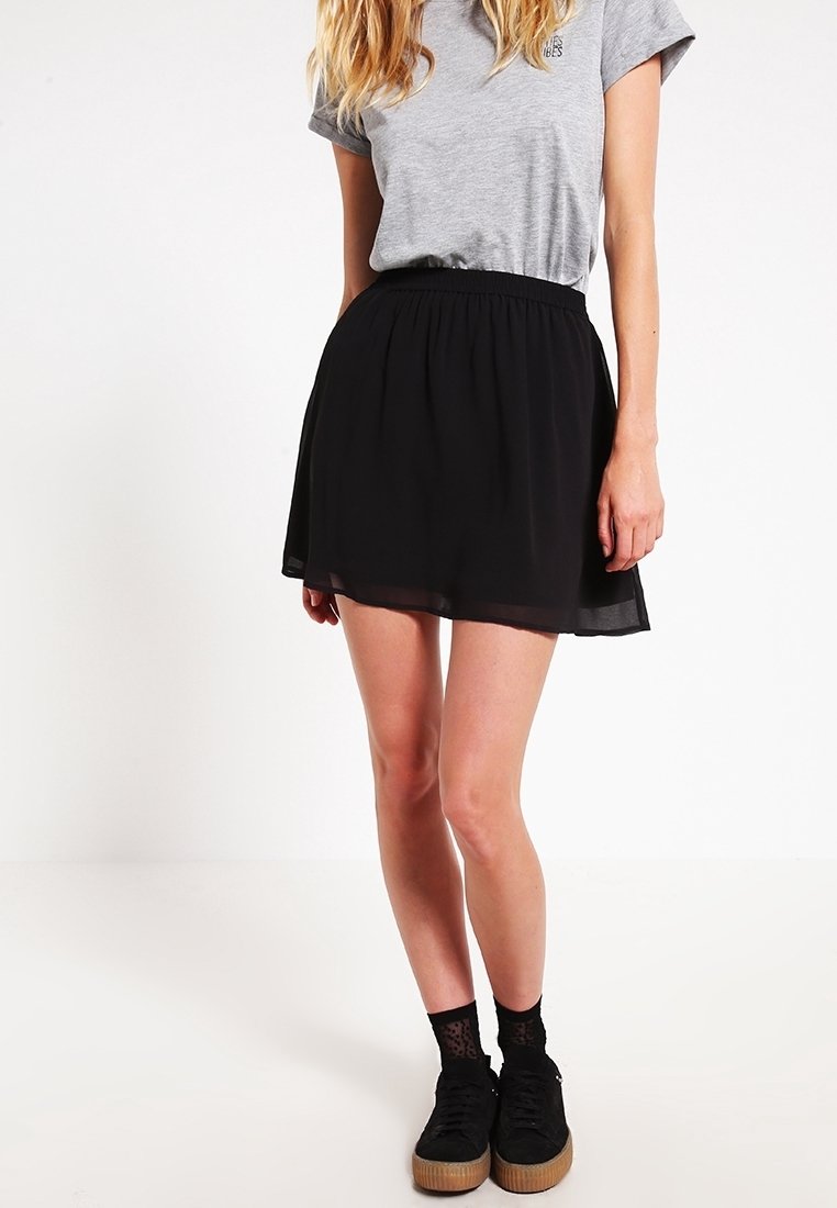 Femme portant un t-shirt gris rentré dans une jupe noire aux genoux, des chaussettes noires à motifs et des baskets à plateforme noires.