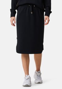 Zwarte midi-rok van zachte stof, met een elastische tailleband met trekkoord en een zijsplits. Gekleed met witte sportschoenen.