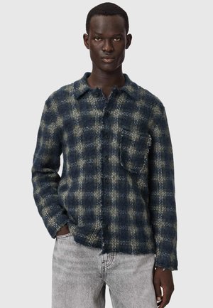 Homme portant une chemise à carreaux bleue et grise avec une poche poitrine et un jean gris clair, debout avec une main dans la poche.