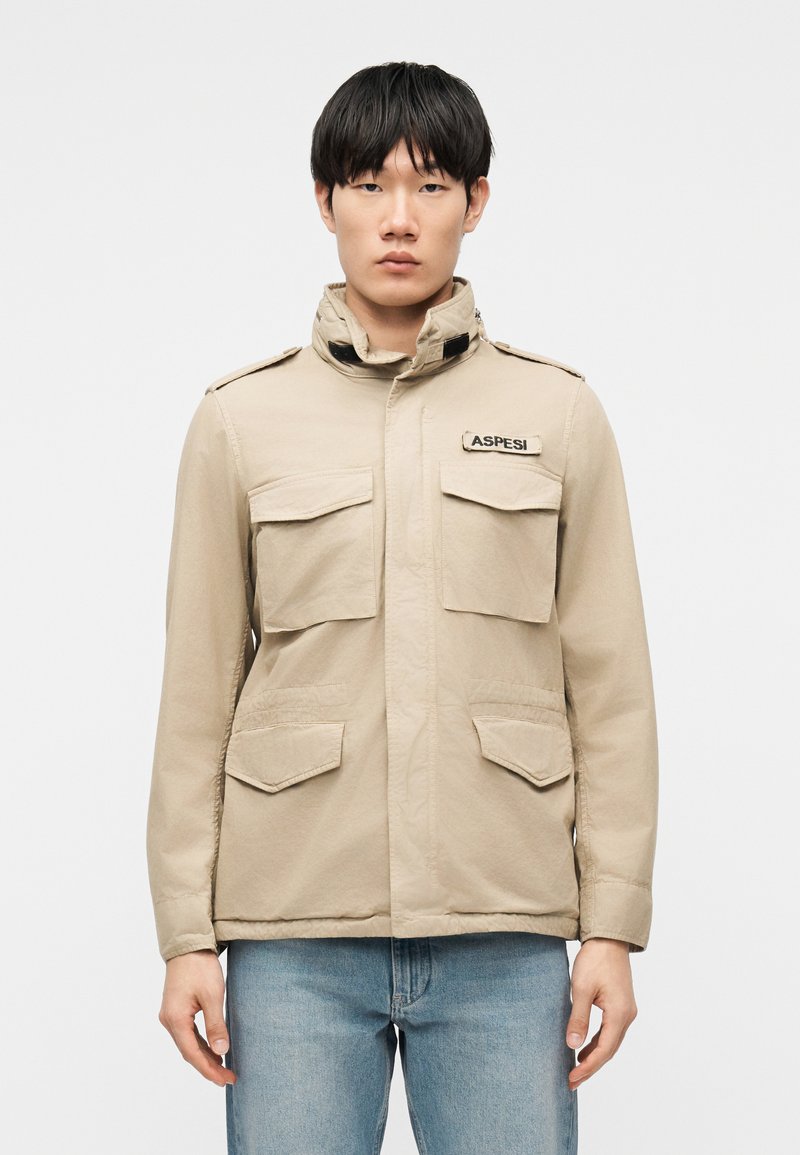 Jeune homme portant une veste beige avec quatre poches avant et un patch "ASPESI", associée à un jean bleu clair, sur un fond uni.