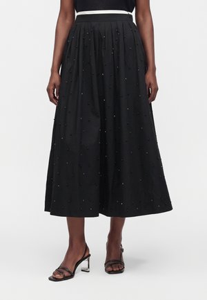 Jupe midi noire avec petites perles décoratives, ceinture blanche, portée avec des talons noirs à brides et talons blocs transparents.