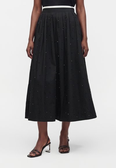 Jupe midi noire avec petites perles décoratives, ceinture blanche, portée avec des talons noirs à brides et talons blocs transparents.