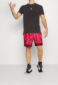 Svart t-shirt med vitt logga, röda mönstrade shorts med zigzagdesign, och svart och vit sneakers. Stående med en hand upplyft.