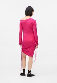 Γυναίκα που στέκεται με πλάτη, φορώντας ένα εφαρμοστό ροζ φόρεμα μακρυμάνικο με μαζεμένες πλευρές και μαύρες μπότες έως τον αστράγαλο με λουριά.