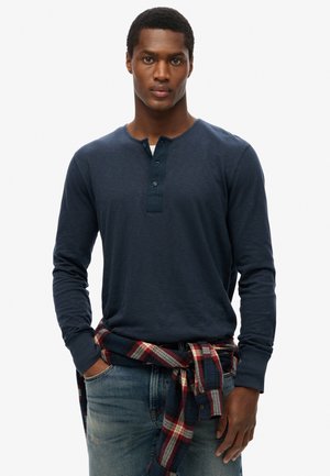 Camisa henley de manga larga en color azul navy con una tapeta de botones, combinada con unos jeans azules y una camisa de franela a cuadros rojo y azul navy atada a la cintura.