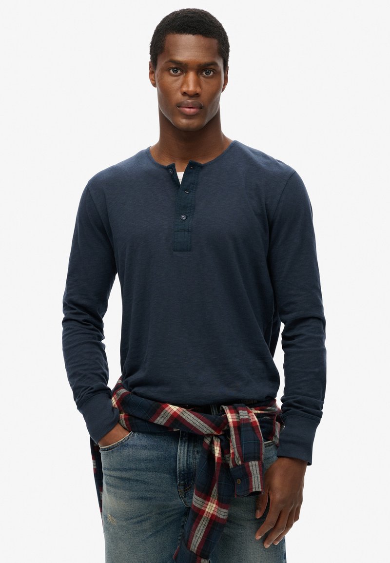 Marineblå langærmet henley skjorte med knaplukning, båret med blå jeans og et rødt og marineblåt ternet flannel skjorte bundet om taljen.