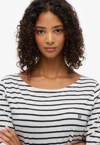 Gestreepte lange mouwen shirt in lichtgrijs met zwarte strepen. Heeft een ronde halslijn en een klein geborduurd logo aan de linkerkant.