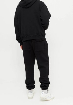 Person iført sort hættetrøje og sorte joggingbukser med baglommer, hvide sokker og hvide sneakers, stående foran en ensfarvet lys baggrund.