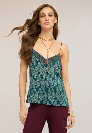 Mujer con cabello largo y castaño lleva una camiseta de tirantes verde con estampado y pantalones granates con textura, complementados con un collar en capas y pulseras de oro.