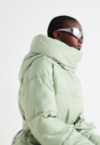 Veste matelassée vert menthe avec col haut et taille ceinturée. Caractérisée par une texture lisse et brillante, elle est accompagnée de lunettes de soleil réfléchissantes.