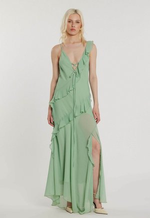 Donna bionda che indossa un abito lungo asimmetrico verde chiaro con volant, una profonda scollatura a V con allacciatura frontale e uno spacco sopra la coscia.