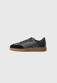 Baskets en cuir noir avec des accents en daim, une semelle en gomme texturée, un bout rond et des lacets noirs, présentant un logo doré sur le côté.