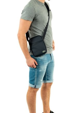 Schwarze synthetische Crossbody-Tasche mit Reißverschlusstasche und grünem Logo, diagonal über den Körper getragen. Glatte Textur und kompaktes Design.