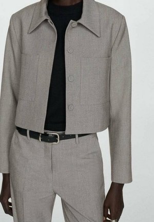 Persoon die een beige cropped colbert draagt met bijpassende broek met hoge taille, een zwarte riem en een zwart topje eronder.