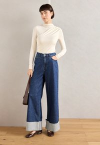 Maglietta a maniche lunghe color crema con dettagli drappeggiati, abbinata a jeans in denim blu a gamba larga con orli grigi sfilacciati e scarpe basse marroni.