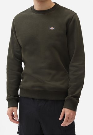 Persona con sudadera de cuello redondo verde oscuro con un pequeño logotipo rectangular en el pecho y pantalones negros, con las manos relajadas a los lados.