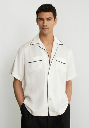 Bărbat cu păr scurt și creț, purtând o cămașă albă cu mâneci scurte cu contur negru și pantaloni negri, mâinile în buzunare, fundal neutru.