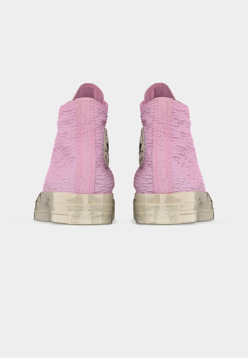 Sneakers alte rosa con texture, suole di gomma bianche consumate e logo Converse All Star sul retro, viste da dietro.