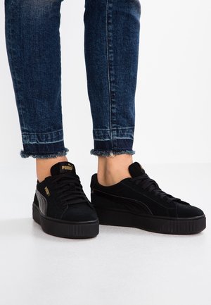 Person trägt schwarze Puma Plateau-Sneaker und dunkelblaue Skinny-Jeans mit ausgefransten Säumen und steht auf einem weißen Boden.