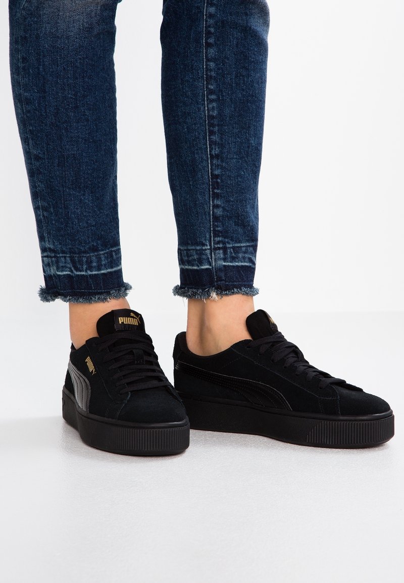 Personne portant des baskets plateformes Puma noires et un jean skinny bleu foncé aux ourlets effilochés, debout sur un sol blanc.