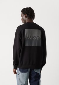 Svart sweatshirt med rund hals, med et stort, glinsende rektangulært grafikk og ordet "HUGO" i en kontrastfarge.