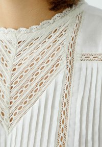 White blouse featuring intricate lace detailing along the neckline and pleated fabric, showcasing a textured pattern with openwork accents.