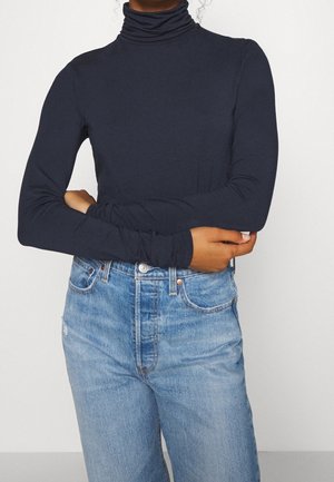 Personne portant un pull à col roulé bleu marine à manches longues et un jean taille haute bleu clair, les bras croisés sur le torse.