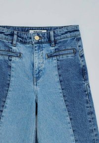 Jeans de mezclilla con un diseño de patchwork que presenta paneles en azul claro y oscuro. Estilo clásico de cinco bolsillos con un botón dorado y cierre de cremallera.