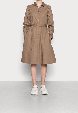 Žena stojící opřená o bílou zeď, oblečená v hnědých košilových šatech s dlouhými rukávy po kolena, s páskem a bílými robustními teniskami.