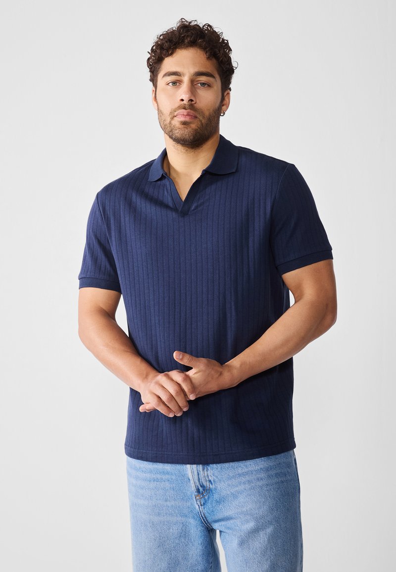Mørkeblå ribbestrikket polo med korte ermer og en myk tekstur. Har flat krage og en avslappet passform. Bæres med lyseblå jeans.