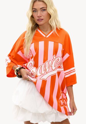 Femme blonde portant un haut oversize rayé orange et rose avec le texte « Bravi », associé à une jupe blanche à plusieurs couches et des boucles d’oreilles créoles dorées.