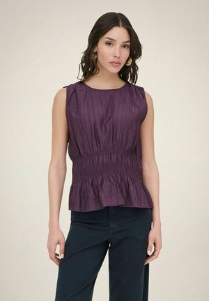 Kobieta o ciemnych włosach, nosząca złote kolczyki-koła, fioletowy top bez rękawów z marszczeniami i ciemne spodnie na jednolitym, jasnym tle.