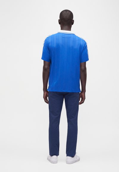 Homme debout, de dos, portant une chemise bleue à manches courtes avec des rayures blanches, pantalon bleu foncé et baskets blanches sur un fond uni.
