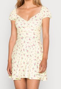 Květinové šaty ve světlé žluté barvě, s nabíranými rukávy, výstřihem do V a přiléhavým korpusem, který se rozšiřuje do volánkové sukně s květinovým vzorem.