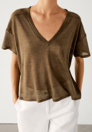 Personne portant un haut marron ample à col en V et manches courtes, légèrement rentré dans un pantalon blanc, les mains dans les poches, sur un fond uni.