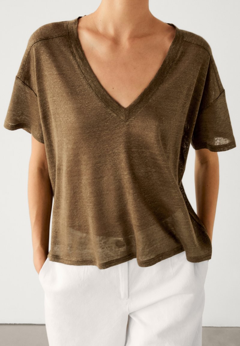 Personne portant un haut marron ample à col en V et manches courtes, légèrement rentré dans un pantalon blanc, les mains dans les poches, sur un fond uni.