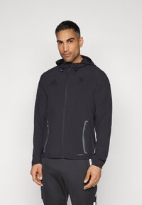 Giacca leggera nera con zip frontale, cappuccio e due tasche laterali con zip. Presenta accenti del logo e polsini elasticizzati per una vestibilità ottimale.