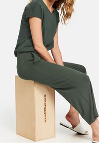Ensemble lounge vert olive comprenant un haut ample et un pantalon large, confectionné dans un tissu doux. Des sandales blanches texturées ajoutent un contraste à l'ensemble.
