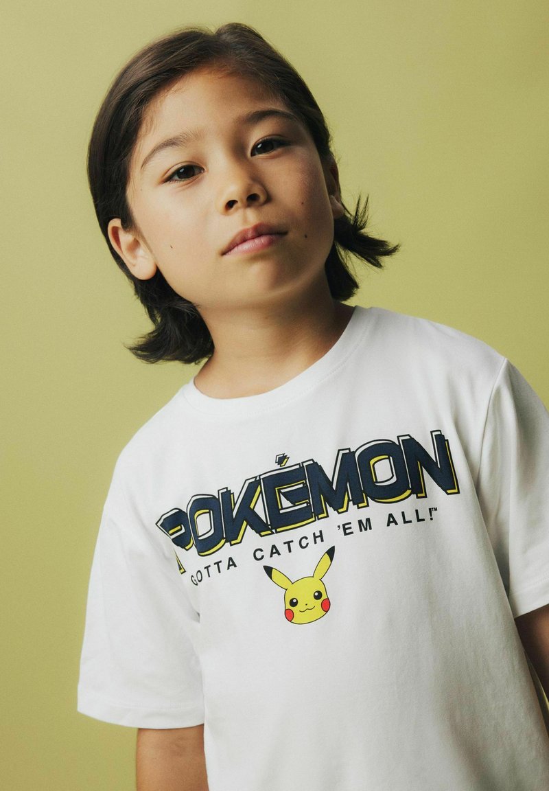 Valge puuvillane T-särk, millel on suur must tekstiprintsi "POKÉMON", kollane Pikachu graafika ja ümmargune kaelus.