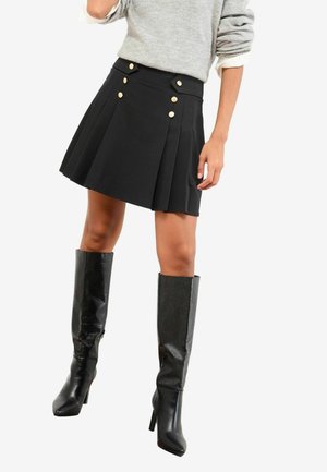 Kvinde iført sort plisseret miniskørt med guldknapper og knæhøje sorte støvler med hæl, kombineret med en grå sweater med opkrullede ærmer.