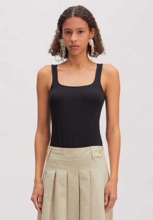 Jonge vrouw met krullend haar draagt een zwarte mouwloze top, een beige geplooide rok met zijknop en hangende pareloorbellen.
