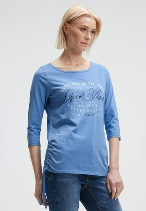 Himmelblaues Langarmshirt mit Rundhalsausschnitt, mit einem Frontdruck und gerafftem Detail auf der linken Seite. Weicher Baumwollstoff.