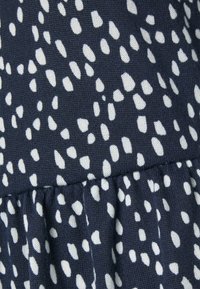 Tecido azul-marinho com manchas brancas irregulares, apresentando uma superfície texturizada e detalhe de volantes franzidos na parte inferior.