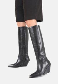 Bottes noires en cuir, montantes jusqu'au genou, avec un embout pointu, un talon compensé, une fermeture éclair sur le côté et une finition élégante. Présente une texture lisse et un detailing minimal.