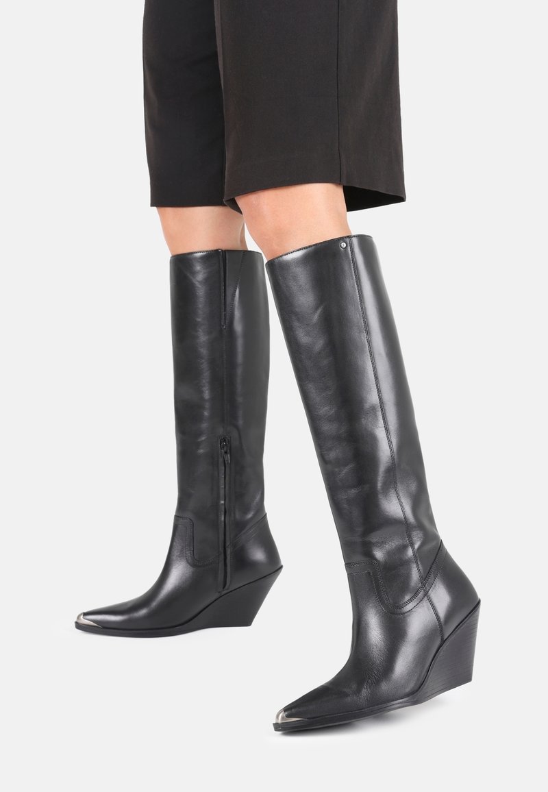 Bottes noires en cuir, montantes jusqu'au genou, avec un embout pointu, un talon compensé, une fermeture éclair sur le côté et une finition élégante. Présente une texture lisse et un detailing minimal.