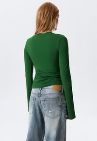 Langärmliges, grünes Oberteil mit taillierter Passform und strukturiertem Muster, kombiniert mit hellblauen, distressed Jeans, die ein braunes Lederetikett tragen.
