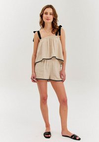 Béžový ľanový overal s vrstveným dizajnom, čiernym vlnitým lemom a nastaviteľnými čiernymi ramienkami. Nosený s čiernymi šľapkami.